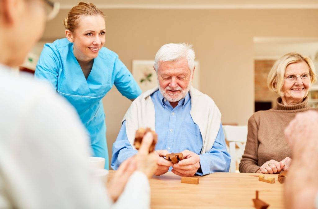 Nurse in blue scrubs standing next to table of seniors playing with a wooden puzzle game.