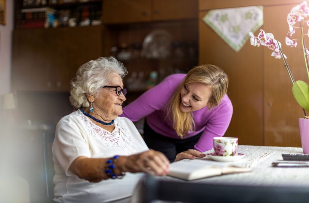 An older adult reading a book while drinking tea. Their adult child is leaning down to look at them with a hand on their shoulder.
