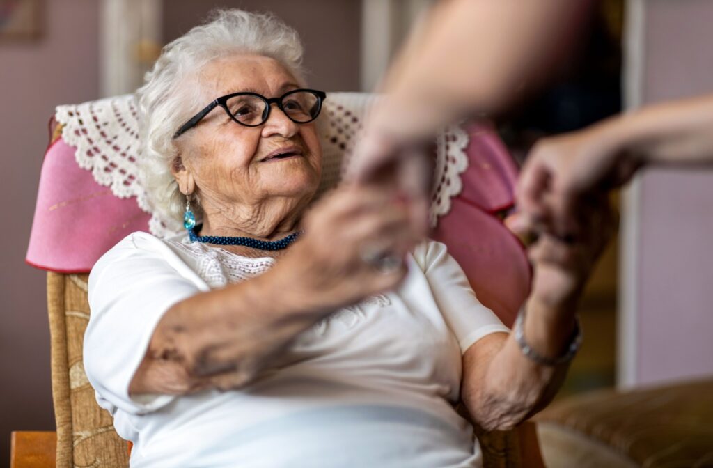 An elderly woman holding her caregiver's hand as they help her stand up from her chair.