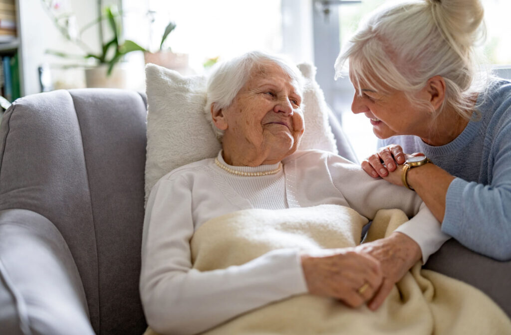 An elderly woman is resting on the couch, while her daughter sits by her side, smiling, as they engage in conversation.