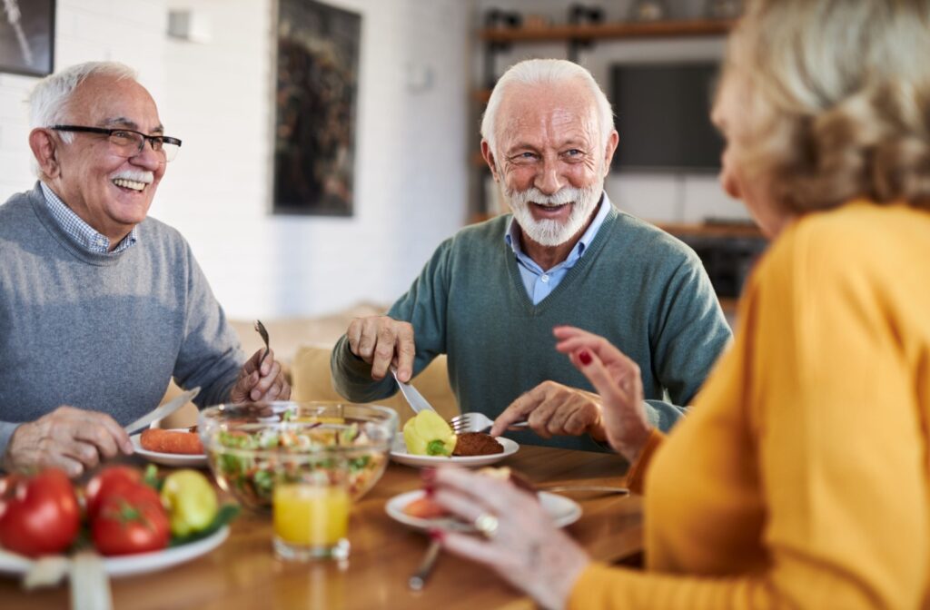 Three older adults smile and talk as they enjoy a colorful meal together in an assisted living community.